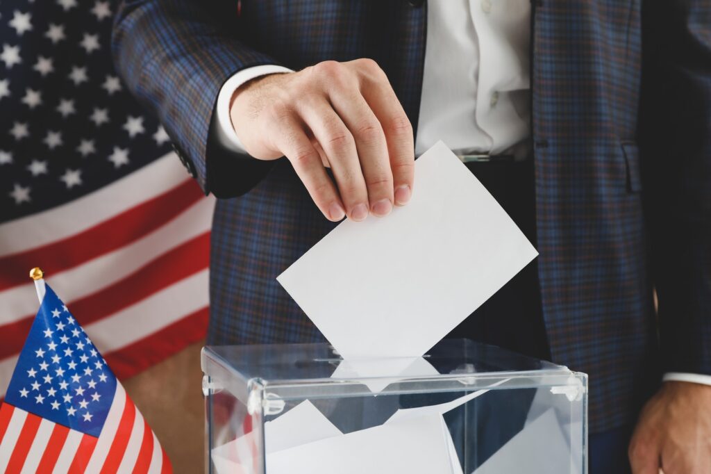 Man putting ballot into voting box against brown background with american flag