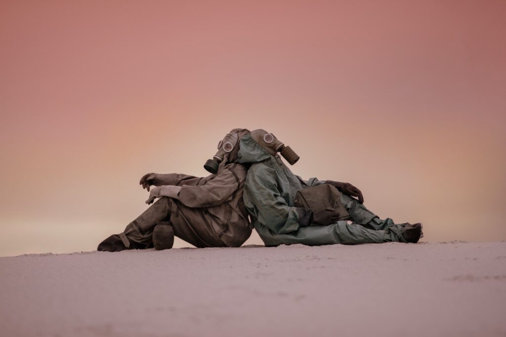 Two men in gas masks and protective suits are sitting on the sand with their backs to each other.