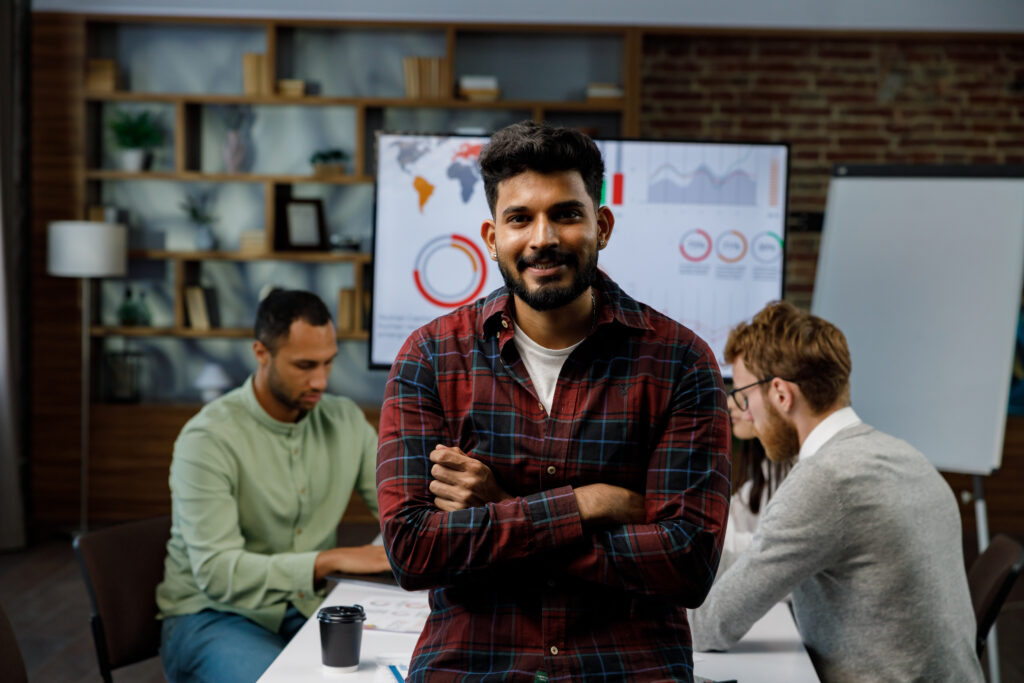 Confident smiling Indian guy, startup team leader, standing in front of mixed race office workers or