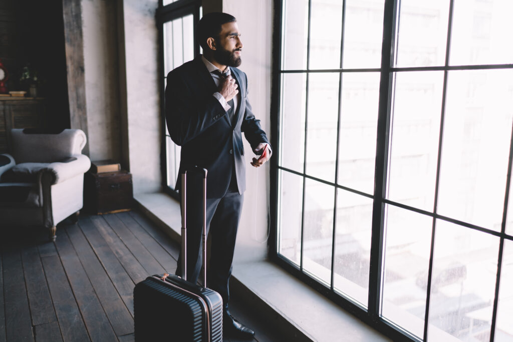 Stylish ethnic businessman standing with suitcase near hotel room window