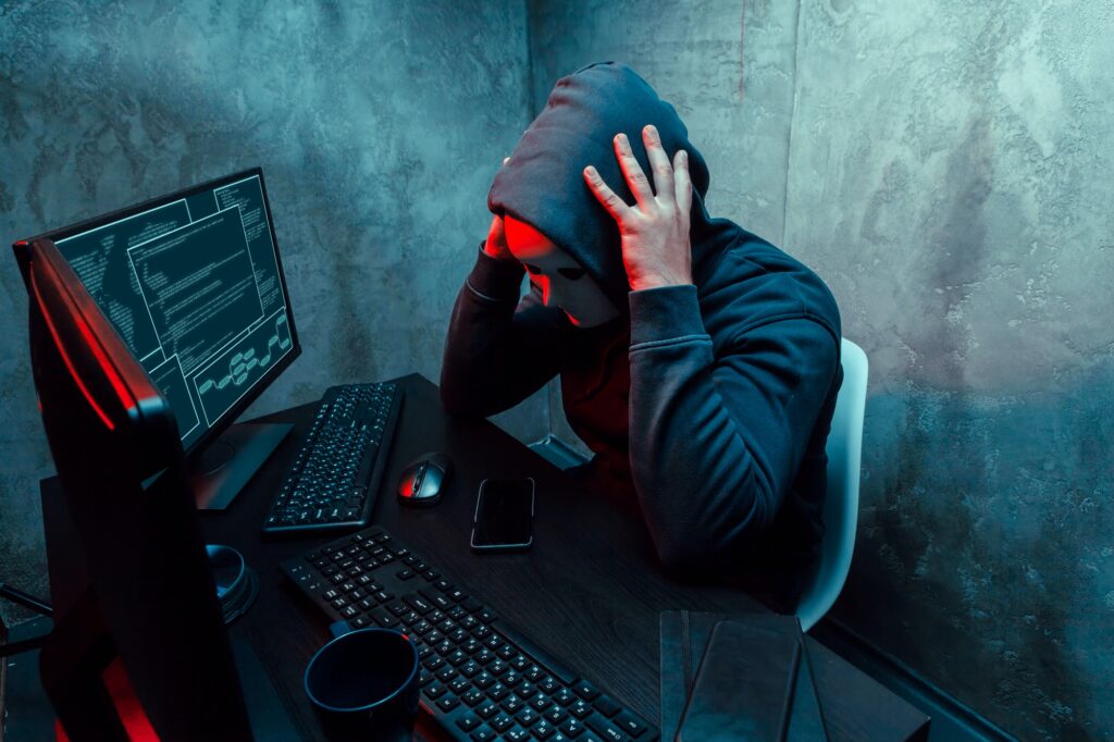 Anonymous internet hacker with mask sitting at the table and using computer