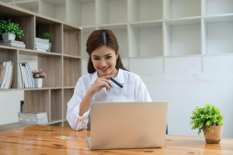 Young adult Asian female consumer holding credit card and laptop at home doing online banking