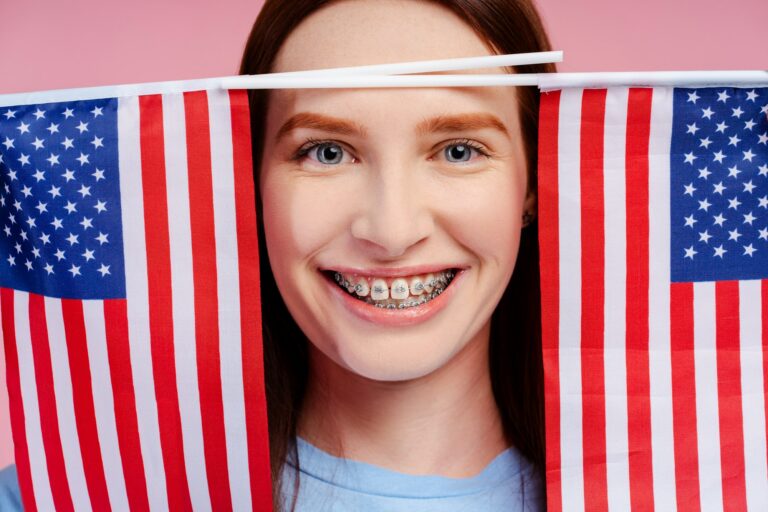 Happy redhead beauty with braces, clutching twin U.S. flags, isolated on a pink background