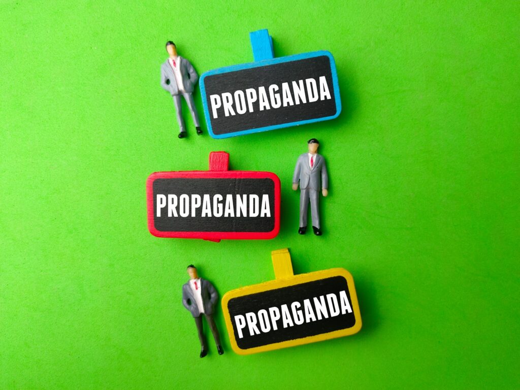 Miniature people and wooden board with the word PROPAGANDA on green background.