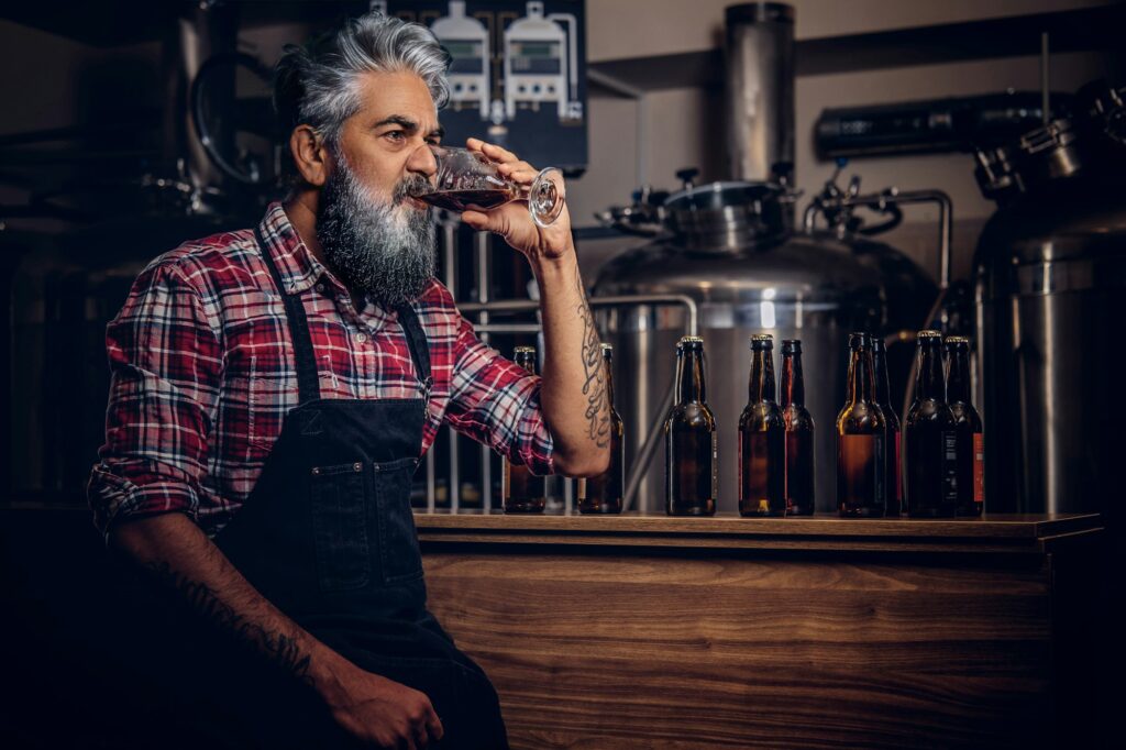 Old man with apron tasting beer in modern brewery