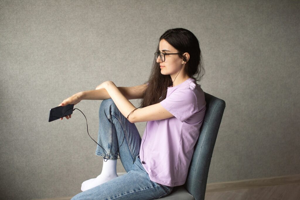 A young girl with glasses and headphones holds a phone in her hands and looks away