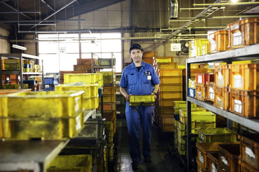 Japanese man wearing baseball cap and blue overall in factory.