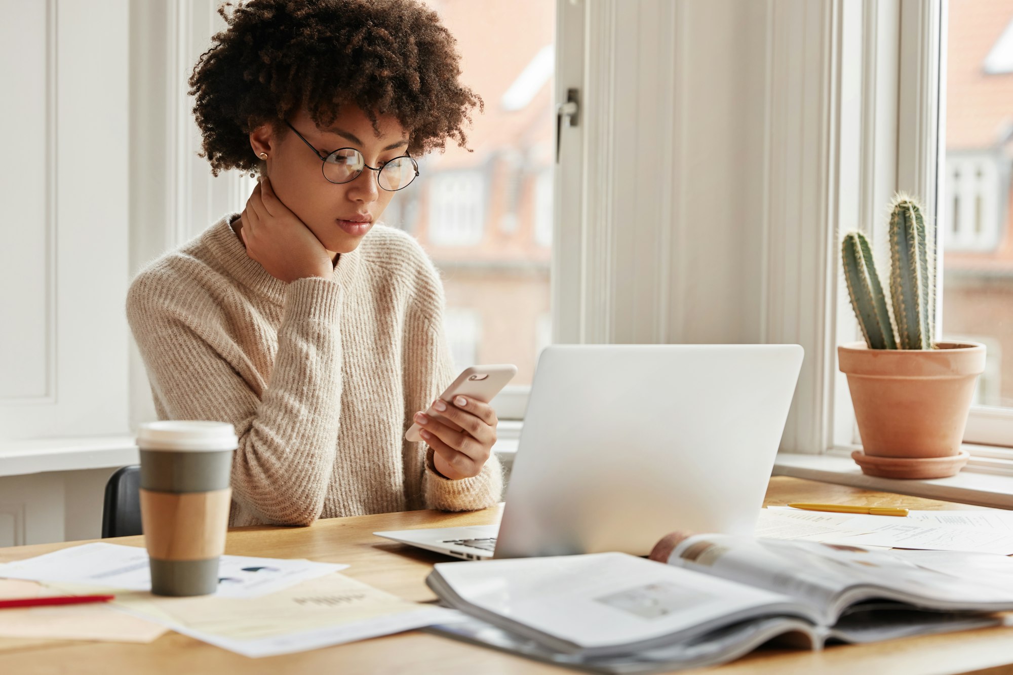 Serious female worker sits at desktop with laptop computer, surrounded with papers and journals, typ