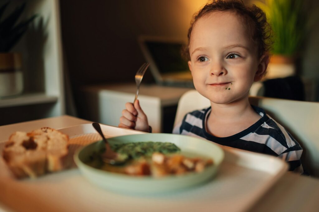 A happy toddler with messy face with fork is eating his food at home.