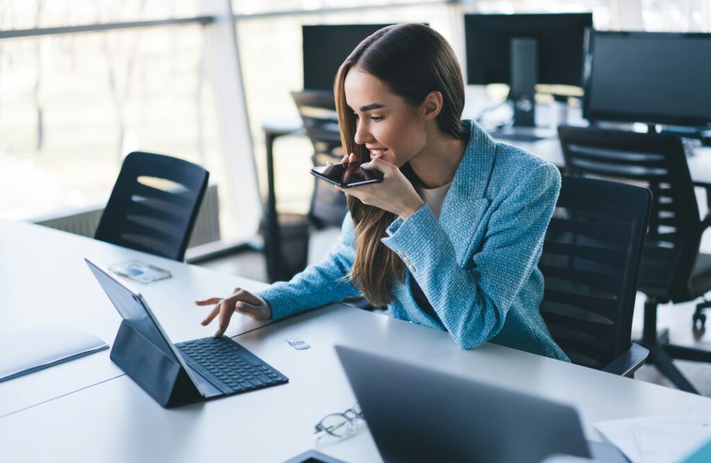 Cheerful young woman working on laptop while holding smartphone