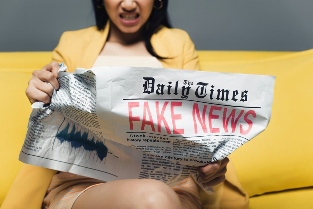 cropped view of angry asian businesswoman sitting on yellow sofa and reading newspaper with fake