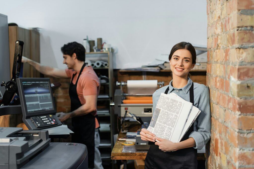 joyful typographer in apron holding newspapers near colleague on blurred background