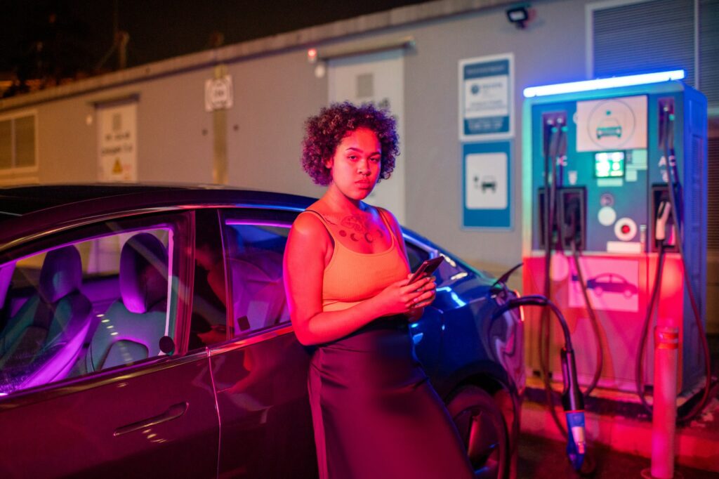 Pretty girl with smartphone standing by electric car