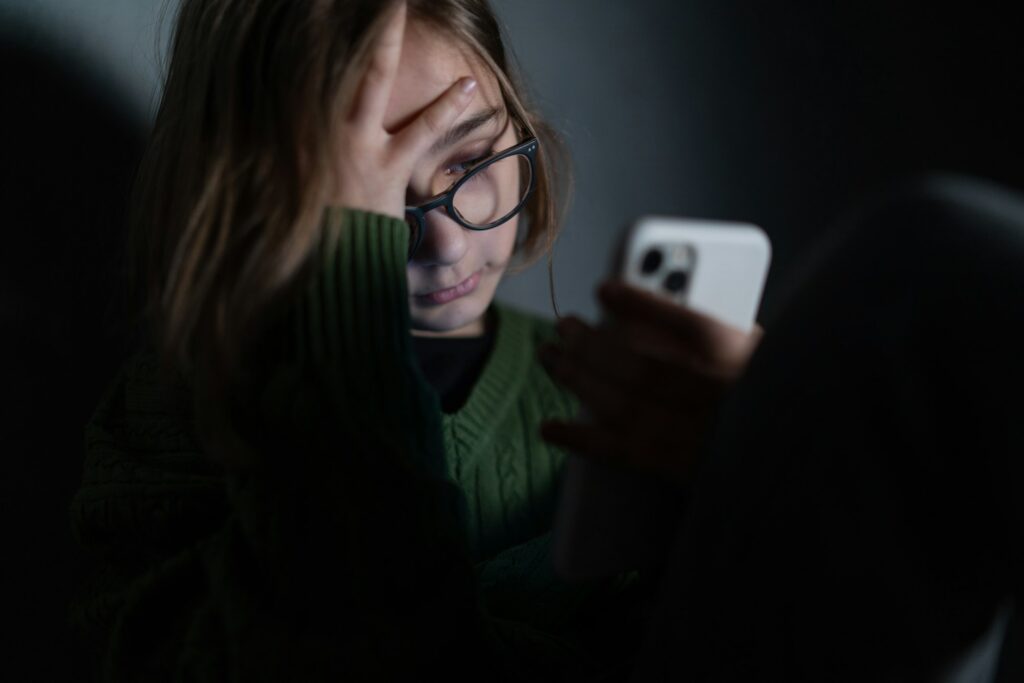 Sad little girl, alone in darkness, sitting and using smartphone