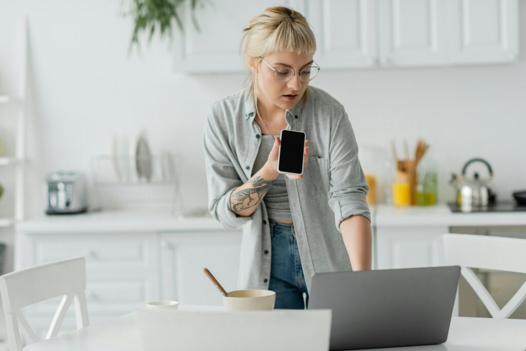 young woman with bangs and tattoo on hand recording voice message on smartphone while standing