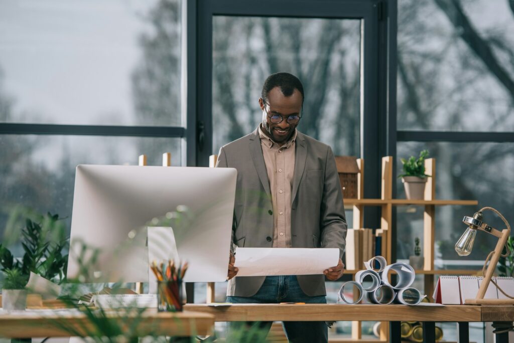 african american architect working with blueprints at workplace with computer