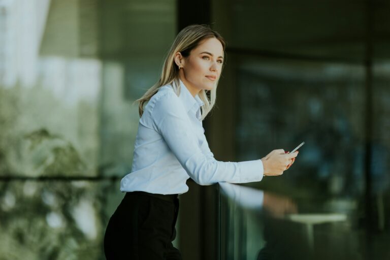 Blonde woman reading smartphone outside modern office building in afternoon shade