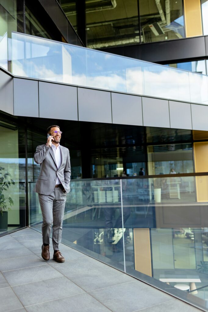 Confident Businessman Smiling in Sharp Suit Against Modern Glass Office Facade. Generative AI