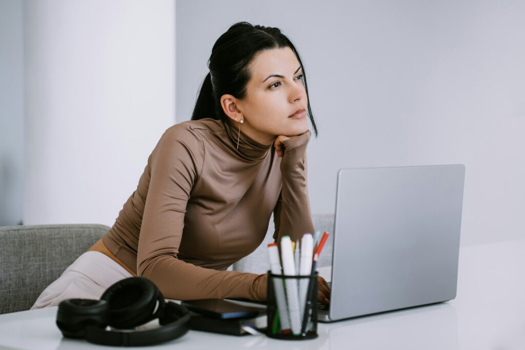 Thoughtful hispanic woman sits at desk with laptop looks away leans on hand, tries find solution