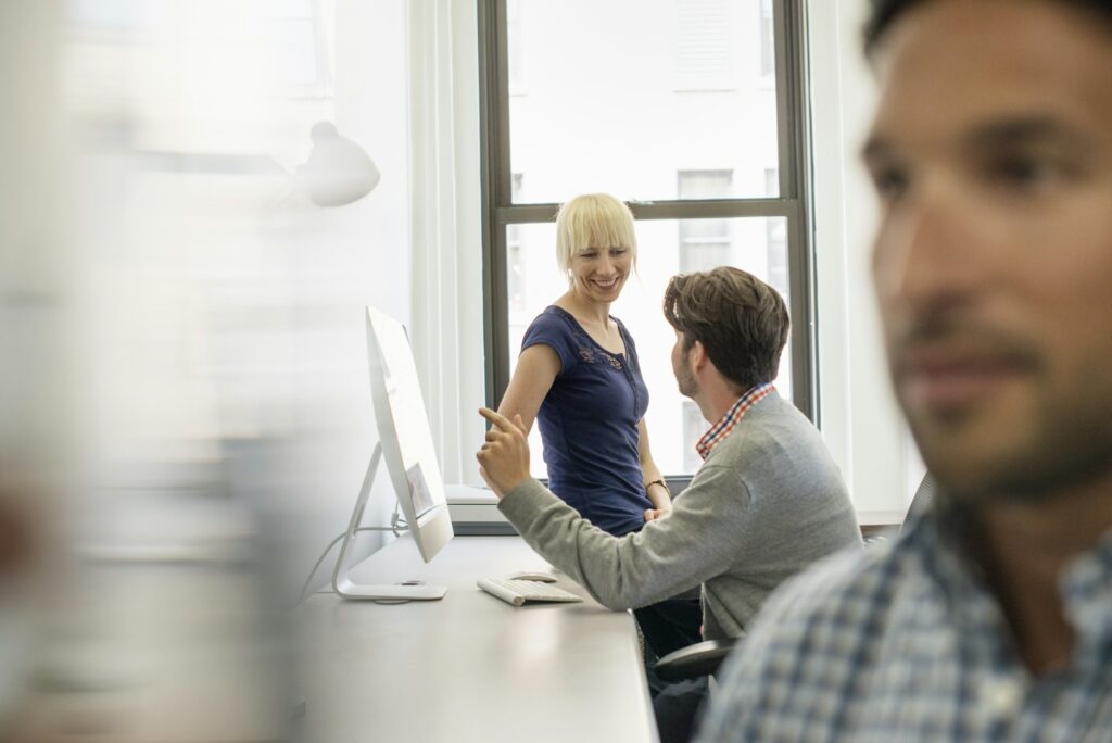 Three business colleagues in an office, two talking and one standing by the door listening.