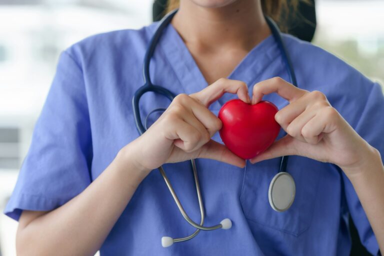 A nurse in blue scrubs holds a red heart in her hands, forming a heart shape around it