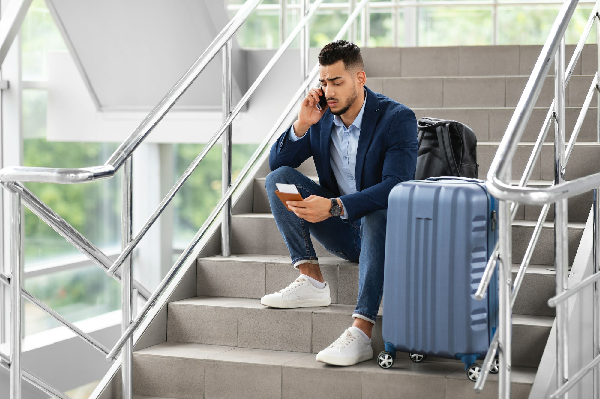 Delayed Flight. Worried Man Sitting On Stairs In Airport, Talking On Cellphone