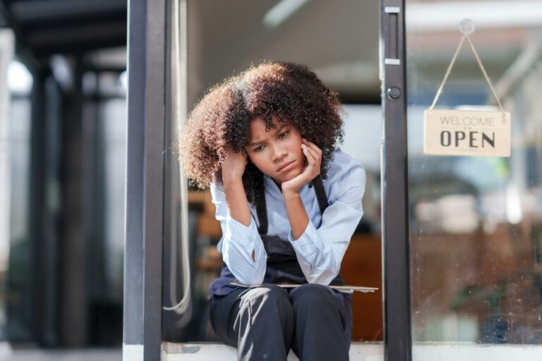 Female shopkeeper sitting stressed out at the store entrance frustrated by the economic impact.