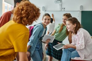 Smiling teen schoolgirls holding digital tablets near blurred friends in classroom in school