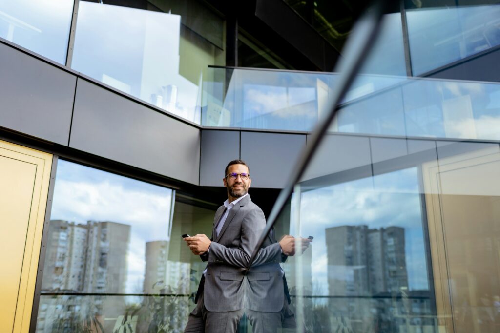 Confident Businessman Smiling in Sharp Suit Against Modern Glass Office Facade. Generative AI