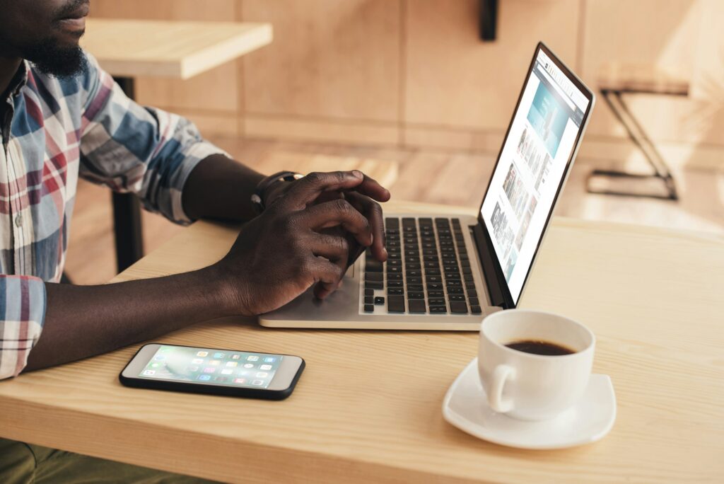 cropped view of african american man using laptop with amazon website and smartphone in coffee shop