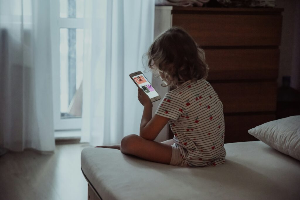 Little blonde girl 4-6 years old sits on the edge of the bed and looks at a mobile phone. Back view.