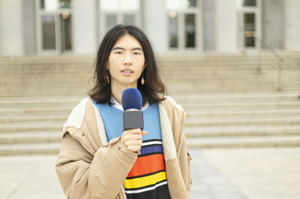 young oriental journalist with a microphone in his hand giving a news in front of a building