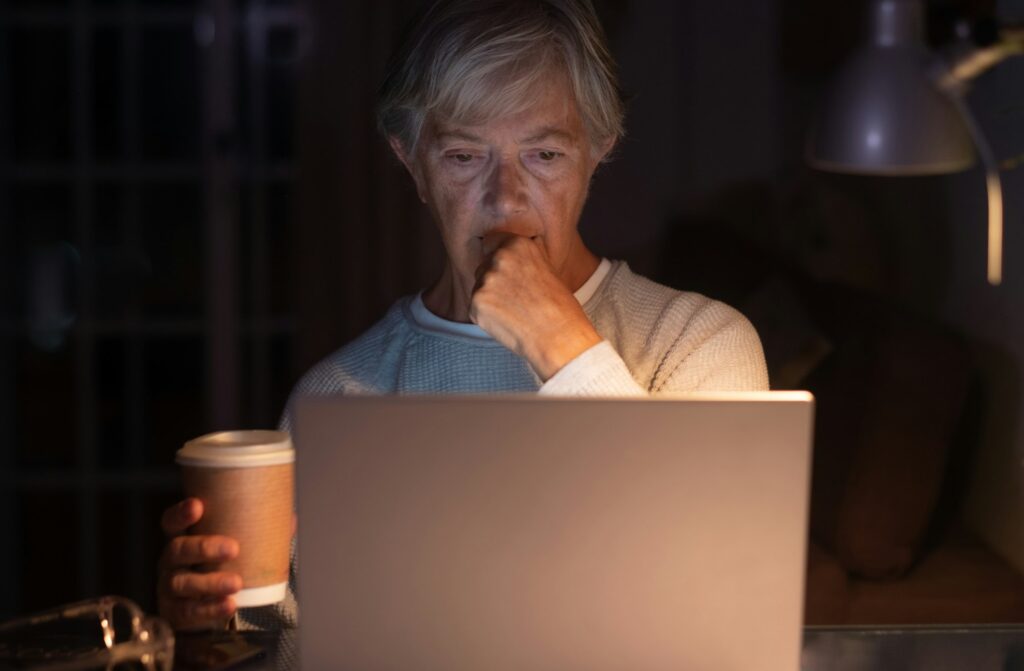 Elderly woman browsing social media content at night using laptop on desk holding a coffee cup