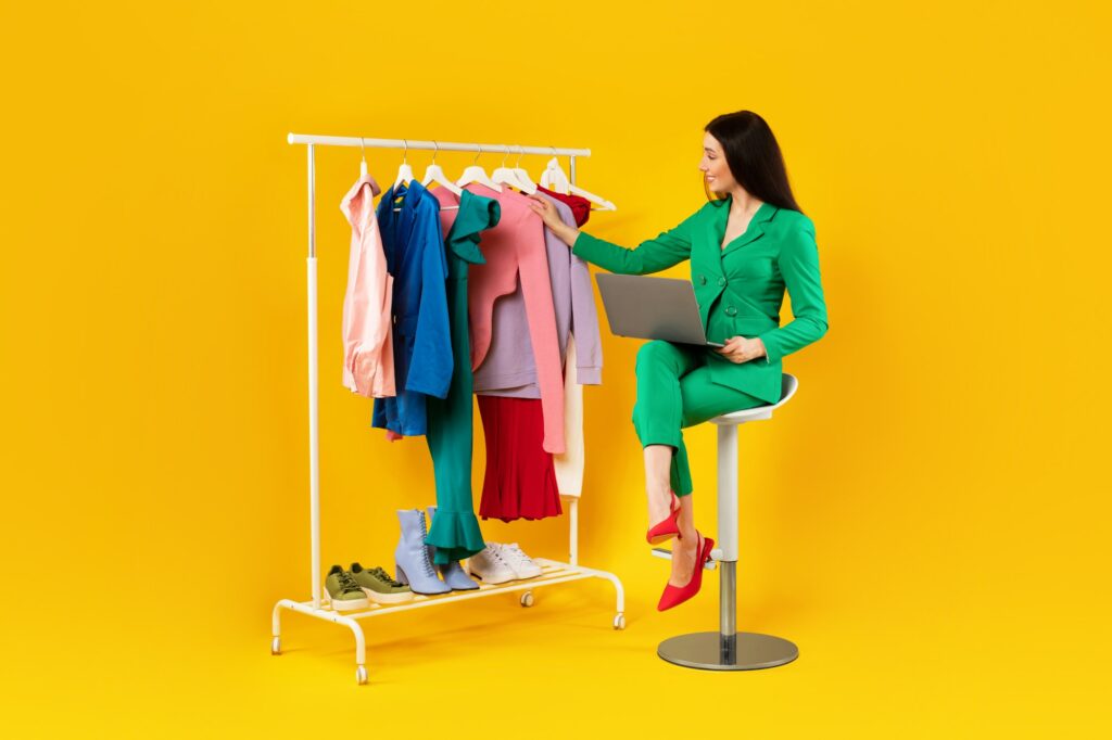 Happy woman fashion designer with laptop looking at clothes on warbrobe rack, using laptop computer