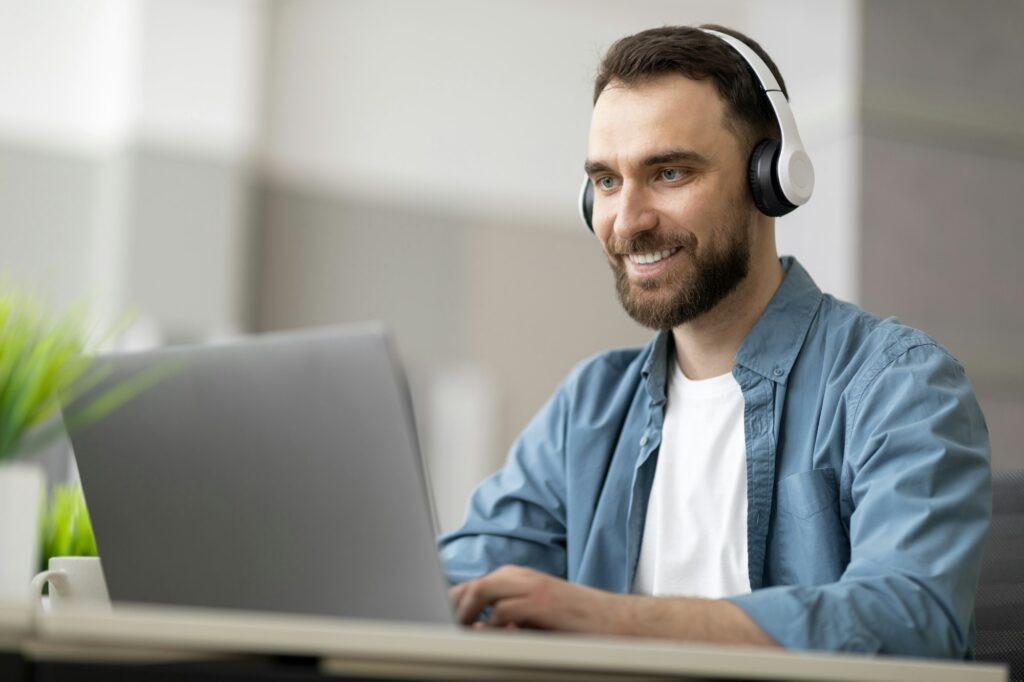 Smiling male employee wearing wireless headphones working on computer in modern office