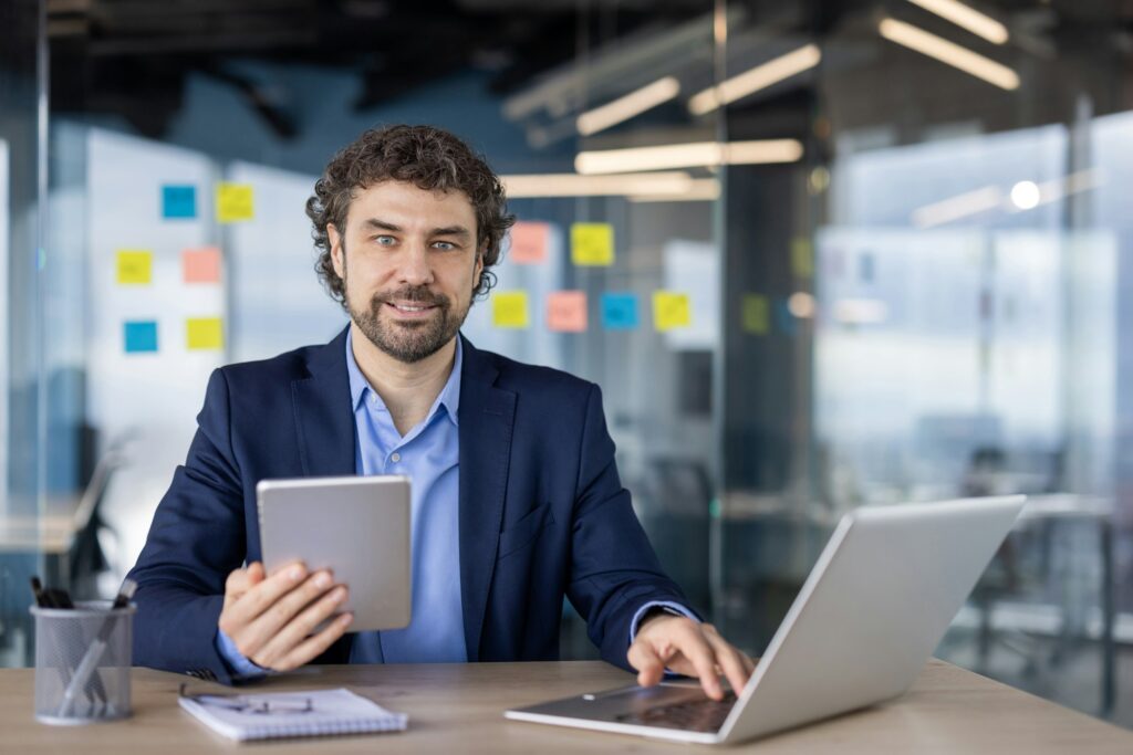 Confident businessman in modern office using tablet and laptop for work productivity and