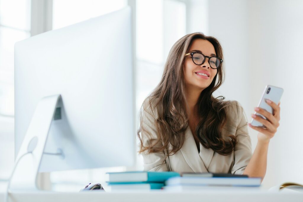 Smiling female in corporate clothes watches funny video via smartphone, sits at desktop in office