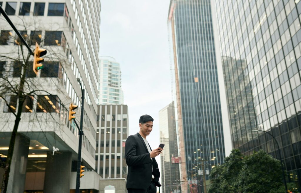 Young happy Asian businessman standing in big city street using phone.