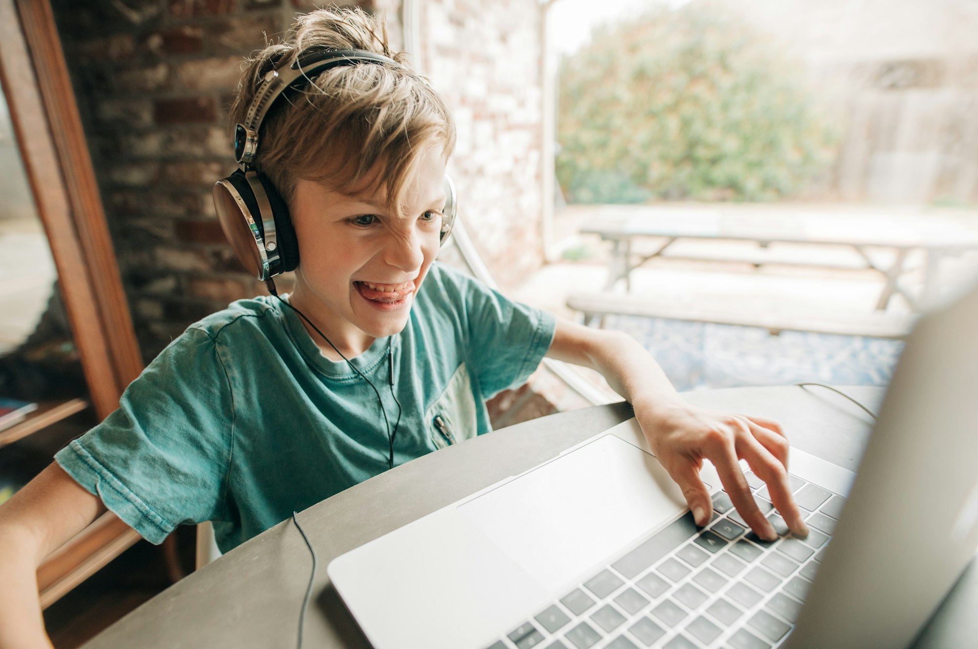 A young gamer playing video games on his laptop device at the table and sticking his tongue out.