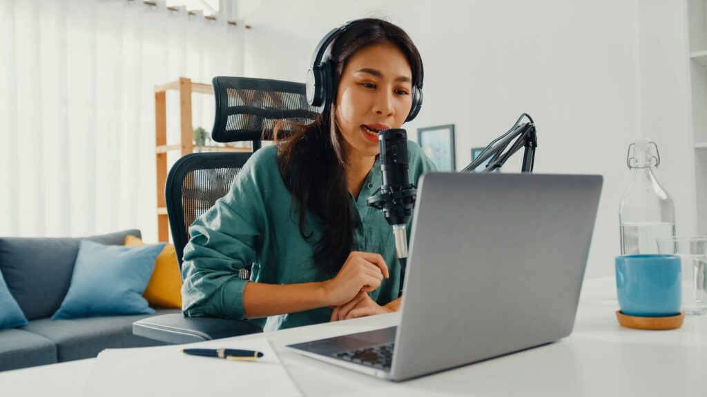 Happy asia girl record a podcast on her laptop computer with headphones and microphone talk.