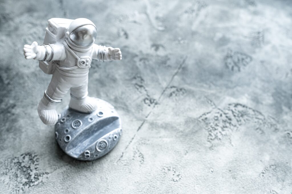 Miniature astronaut toy balancing on moon in spacesuit waving his hand.