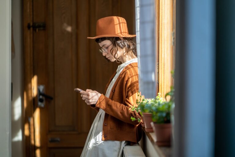 Pensive hipster girl standing with phone near window at home using taxi booking mobile app