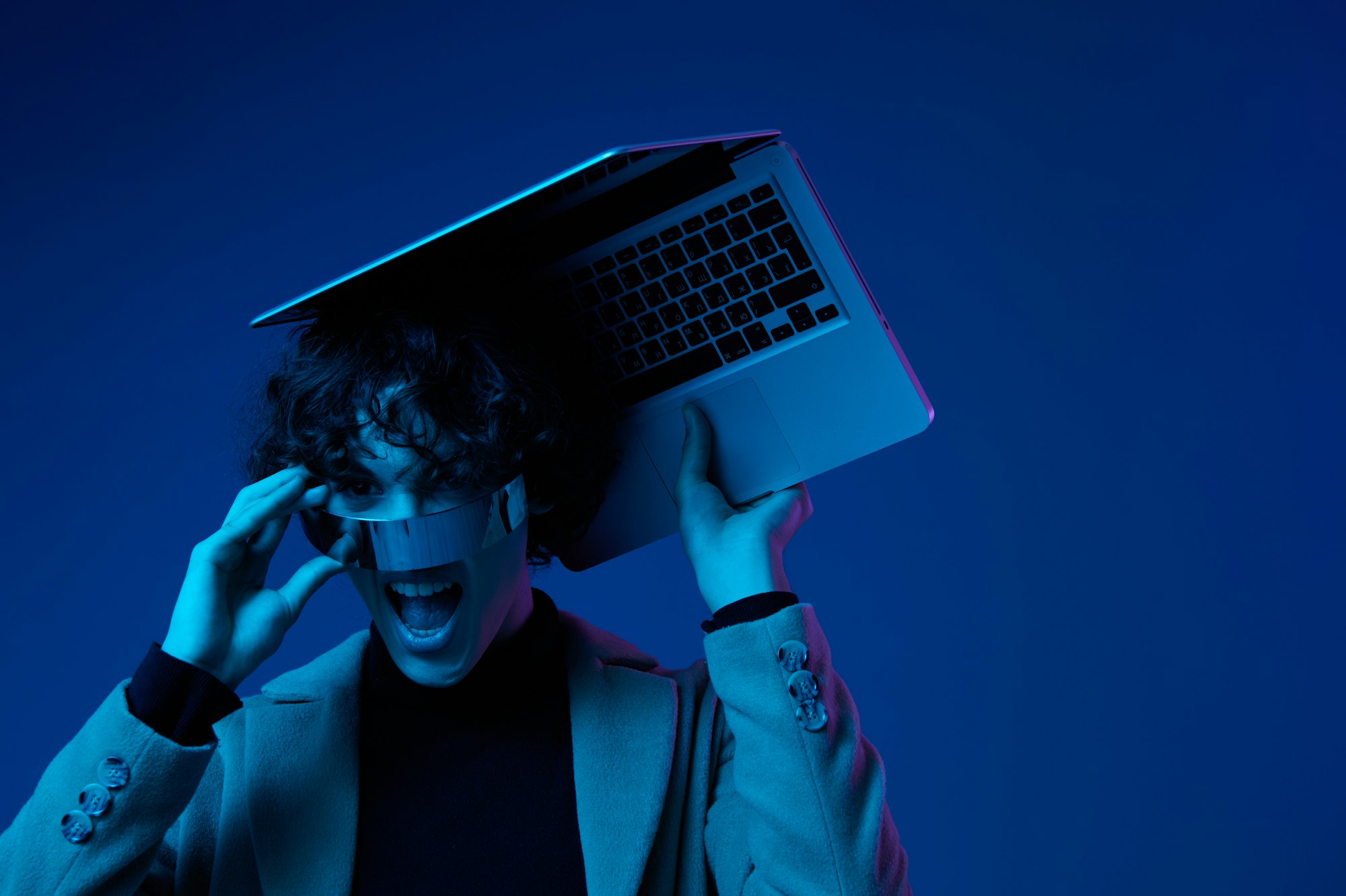 Portrait of stylish man hacker with laptop and futuristic glasses in blue light, cyber security