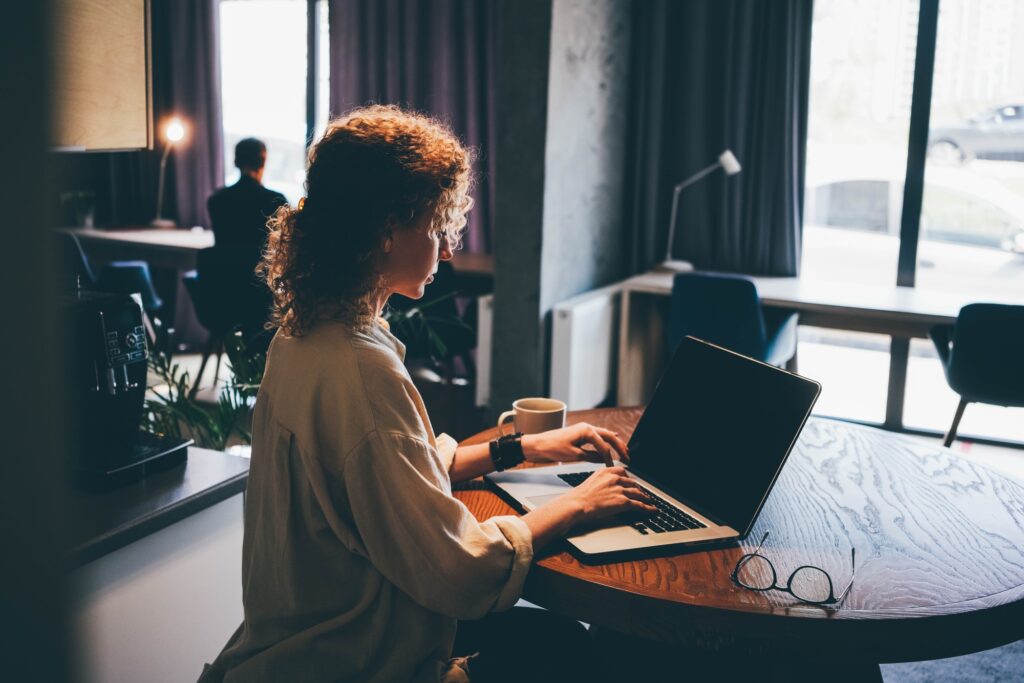 Woman with leather bracelet types on modern laptop keyboard at workplace against blurry colleague