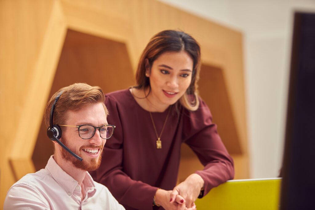 Businessman Wearing Headset Talking To Colleague In Busy Customer Services Centre