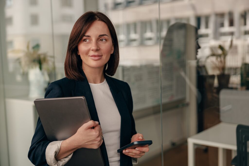 Confident businesswoman in a modern office, holding a laptop and smartphone, radiates efficiency