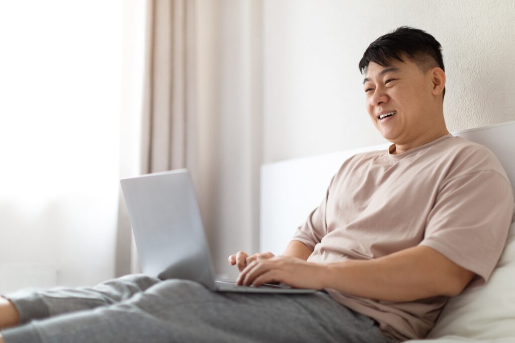 Happy mature asian man using laptop in bed, copy space