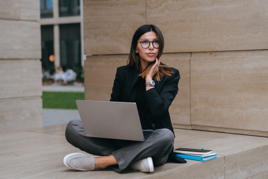 Pensive Hispanic young businesswoman in black jacket using laptop sitting outside looks up dreaming