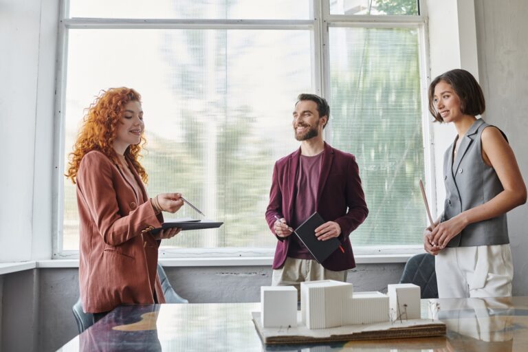 redhead designer talking to business partners near building model, investment in startup project