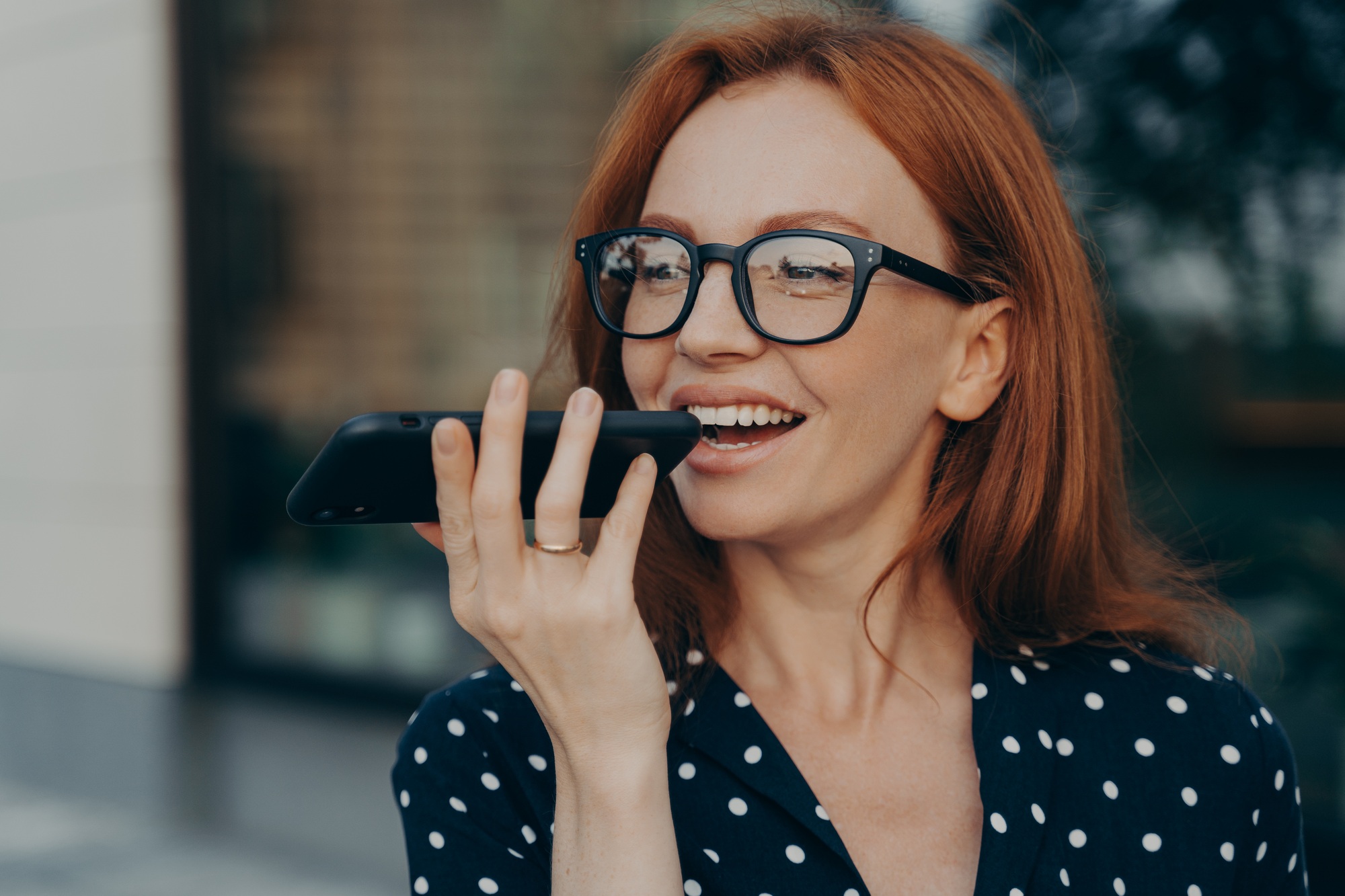 smiling red haired business woman using virtual digital voice assistant while standing outdoors